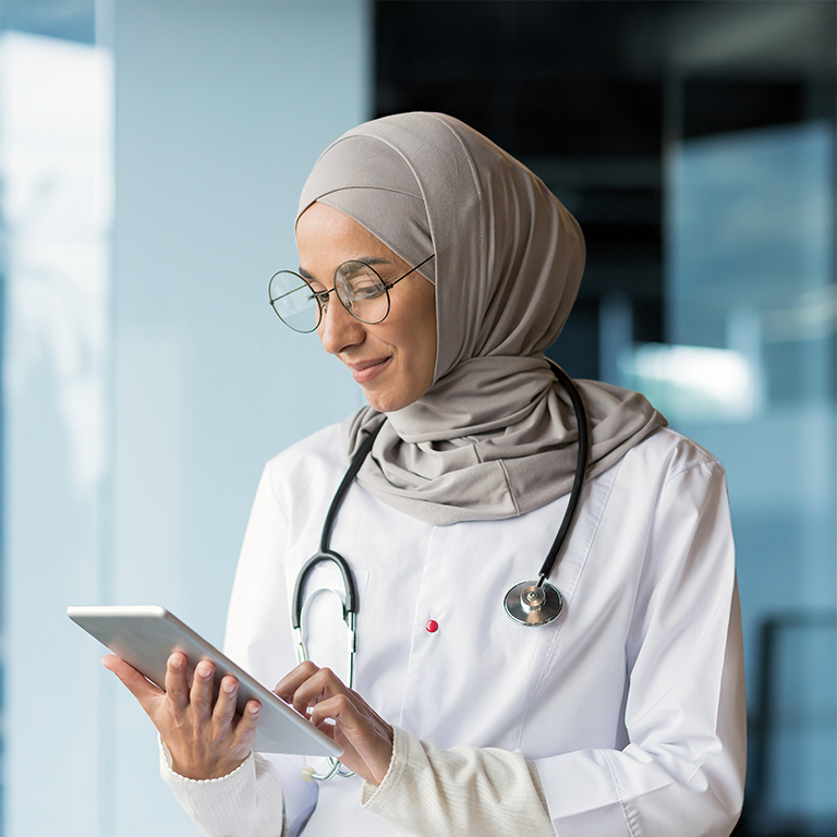 Close-up photo. A young Arab woman doctor is standing in the hospital in the office in a hijab and with a stethoscope. She holds a tablet in her hands, concentrates on working, typing.